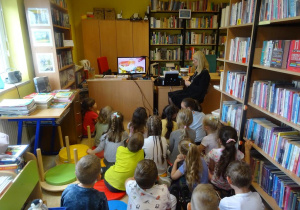 Dzieci siedzą pomiędzy regałami książek, oglądają film edukacyjny.